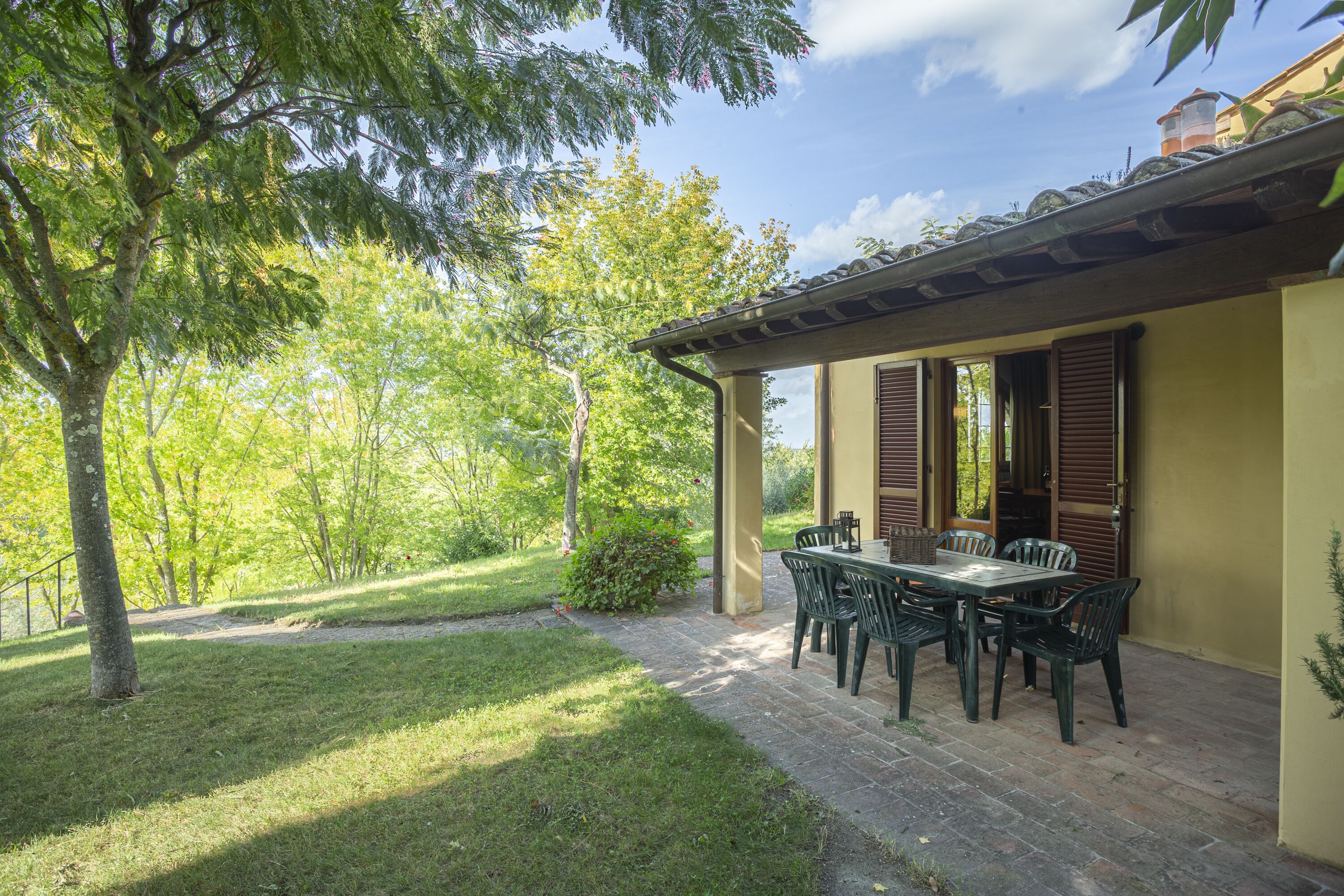 Um pátio com mesa e cadeiras, rodeado por um jardim e um edifício com telhado de azulejos.