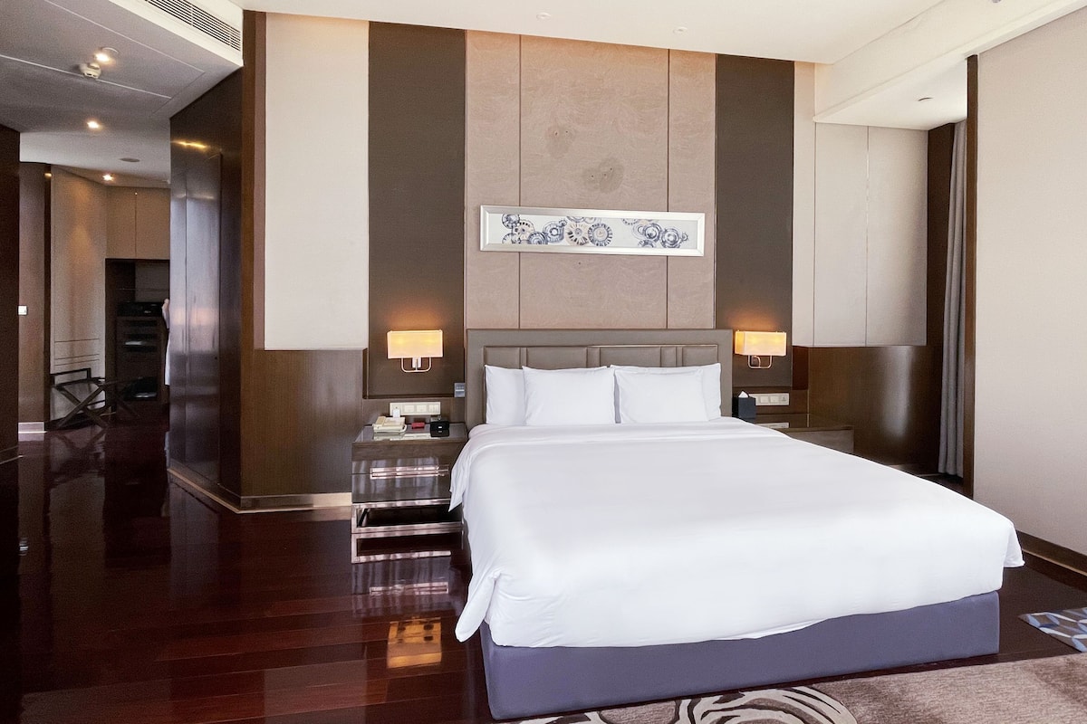 A modern hotel room with a large bed, two bedside tables, a nightstand, a wall-mounted artwork, and a hallway leading to a staircase.