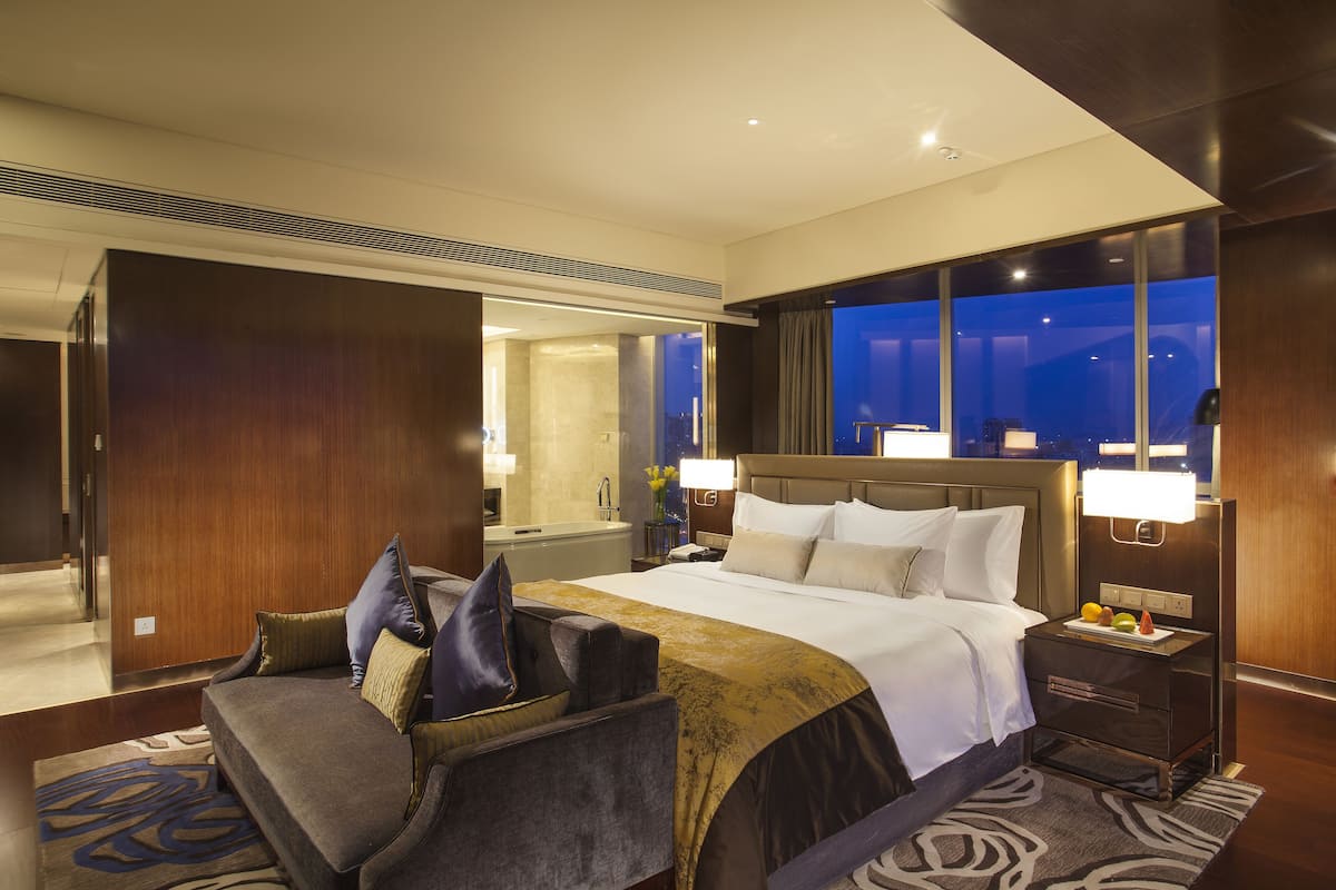 Hotelkamer met een groot bed, een nachtkastje met lamp, een bank en uitzicht op de stad 's nachts.