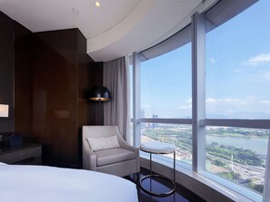 A hotel room with a large window, a bed, a chair, a small table, and a lamp.