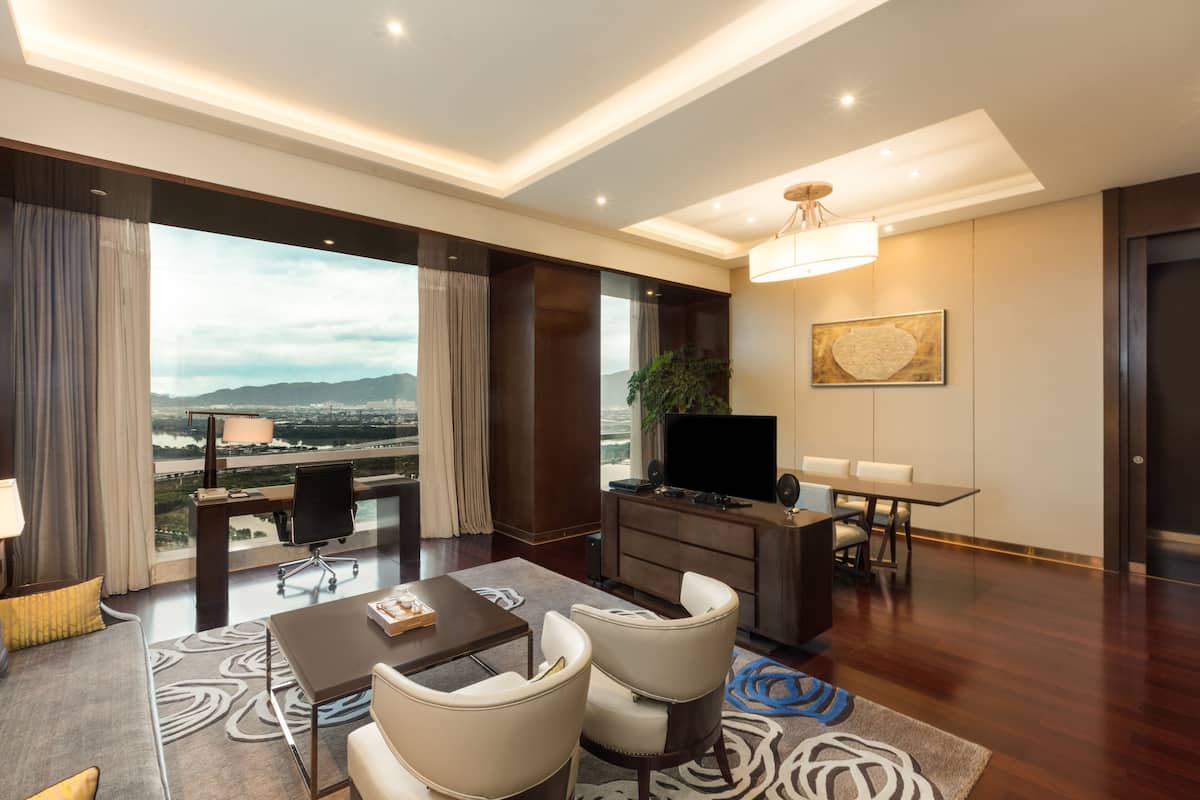 Een moderne hotelkamer met een groot raam dat uitzicht geeft op de stad, een bureau met een flatscreen televisie, een zithoek met stoelen en een bank met een kamerkleed.