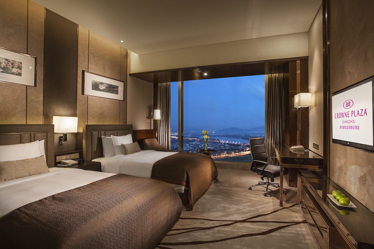 Hotelkamer met twee bedden, een bureau, een stoel, een televisie en een groot raam met uitzicht op de stad.