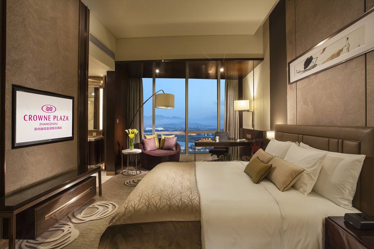 Hotelkamer met een groot bed, een bureau, een stoel, een televisie waarop de naam van het hotel staat, en een uitzicht op de stad door grote ramen.