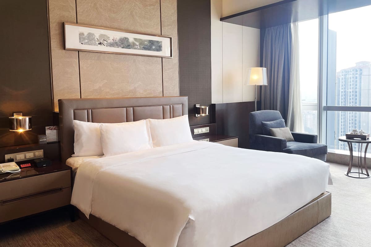 Een hotelkamer met een groot bed, een nachtkastje met telefoon en lamp, een stoel en een raam met uitzicht op de stad.