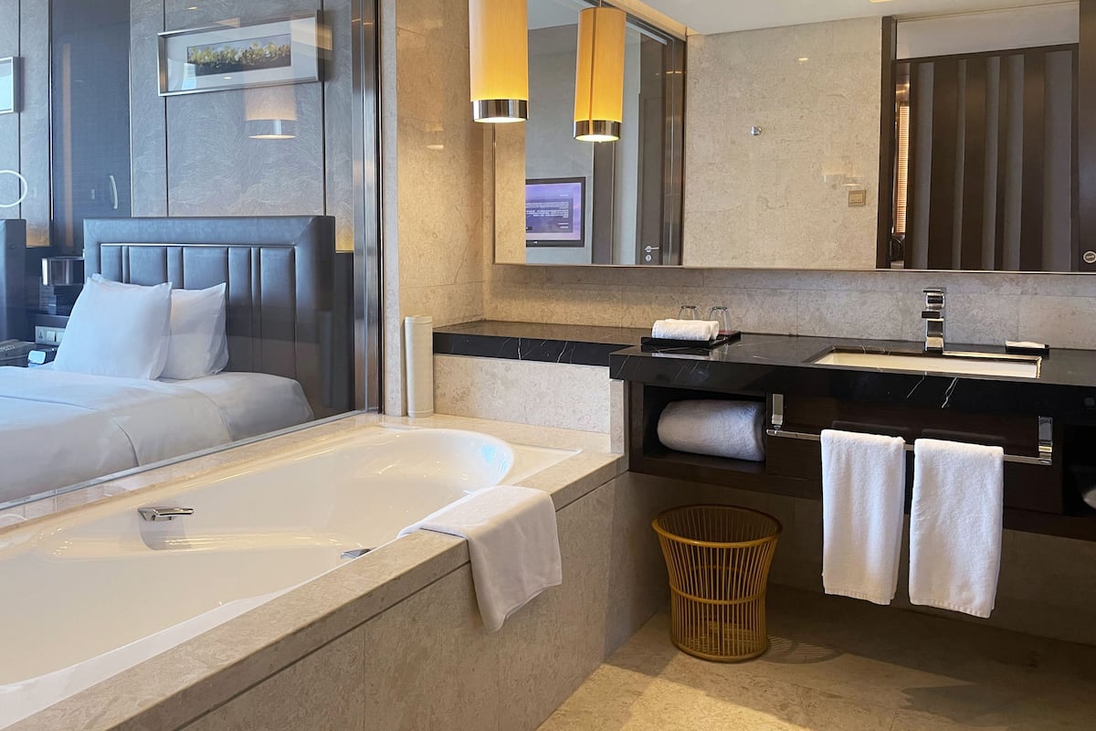 A modern hotel bathroom with a large bathtub, a vanity with a mirror, and a bed with a headboard.
