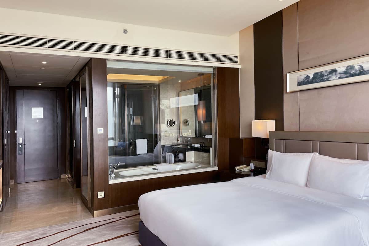 A modern hotel room with a large bed, a glass-enclosed bathroom, and a view of the hallway.