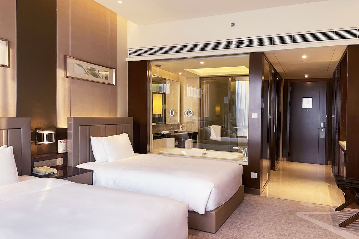 Een hotelkamer met twee bedden, een grote spiegel, een badkamer met een wastafel en spiegel, en een deur naar een andere kamer.