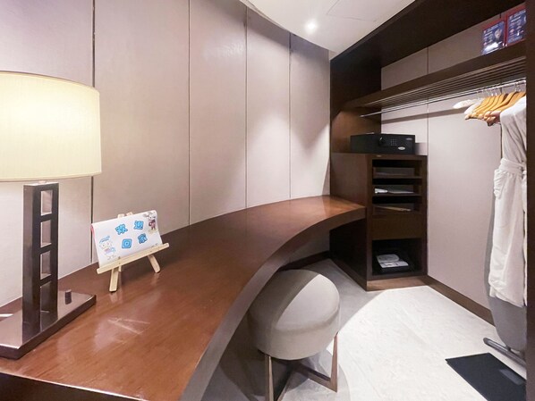 A hotel room with a wooden desk, a small easel with a sign, a chair, a coat rack with a robe, and a shelf with a microwave.