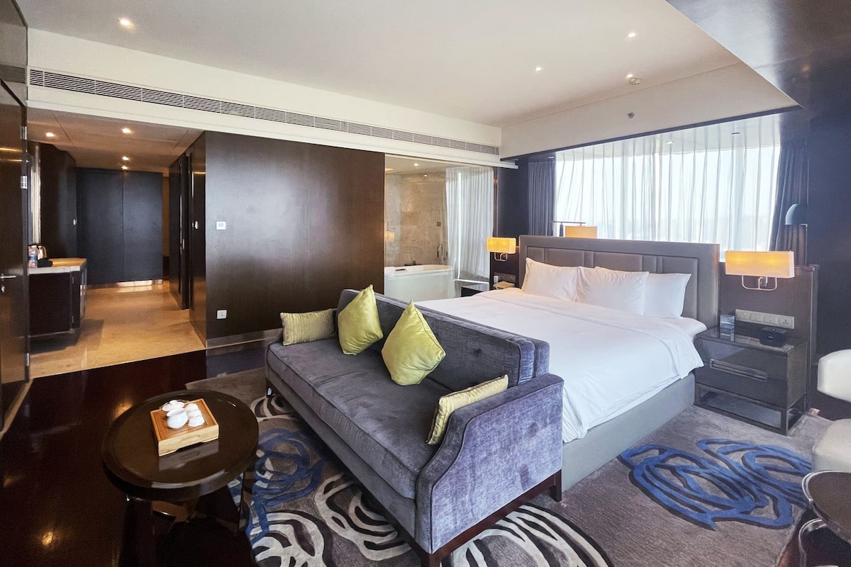 A modern hotel room with a large bed, a sofa, a small table, and a bathroom visible through an open door.