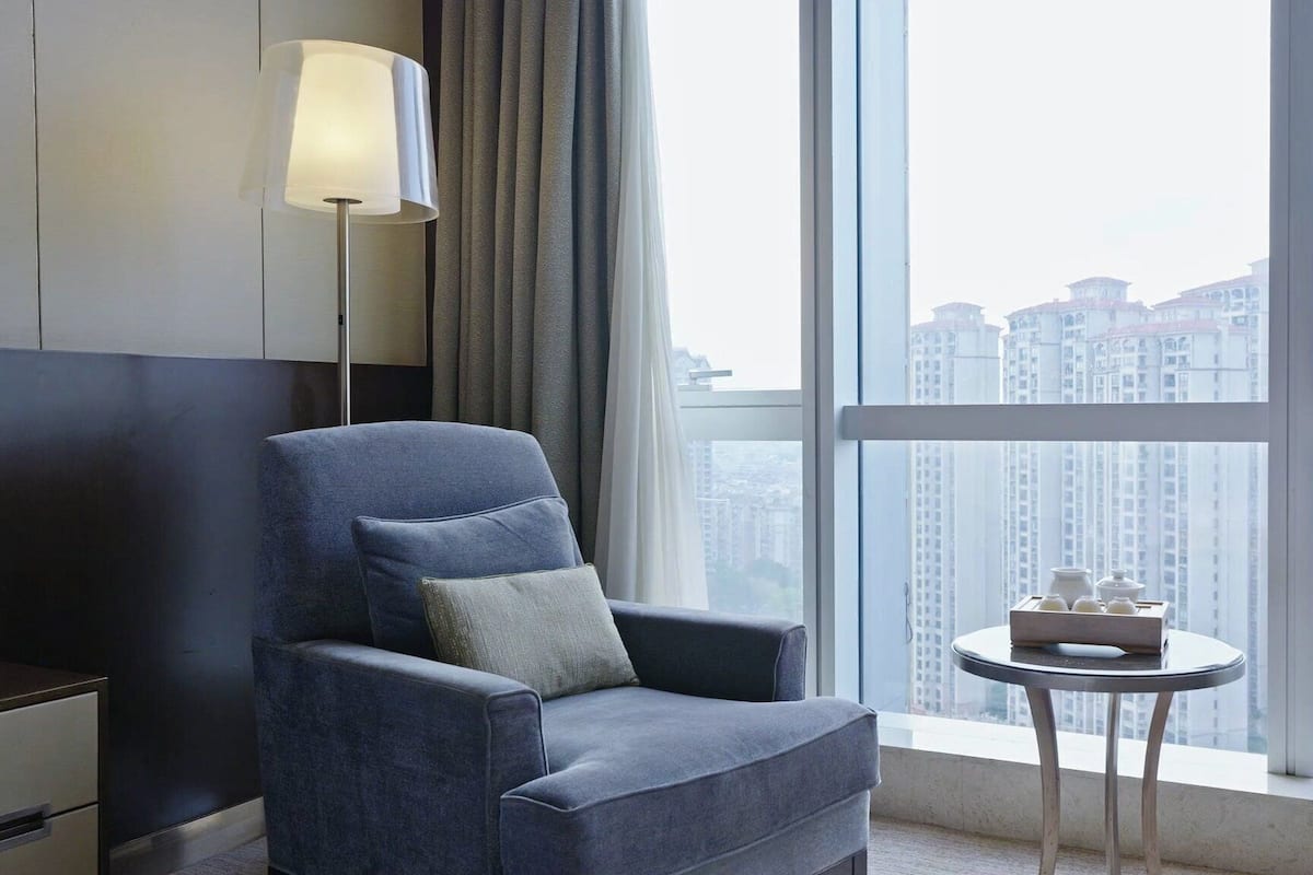 Een moderne hotelkamer met een groot raam, een blauwe fauteuil, een klein rond tafeltje met een dienblad en een lamp aan de muur.