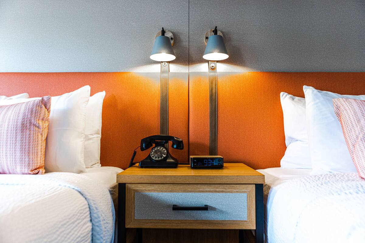 Quarto de hotel com duas camas, uma mesa de cabeceira com telefone e relógio digital, e duas luminárias de parede.