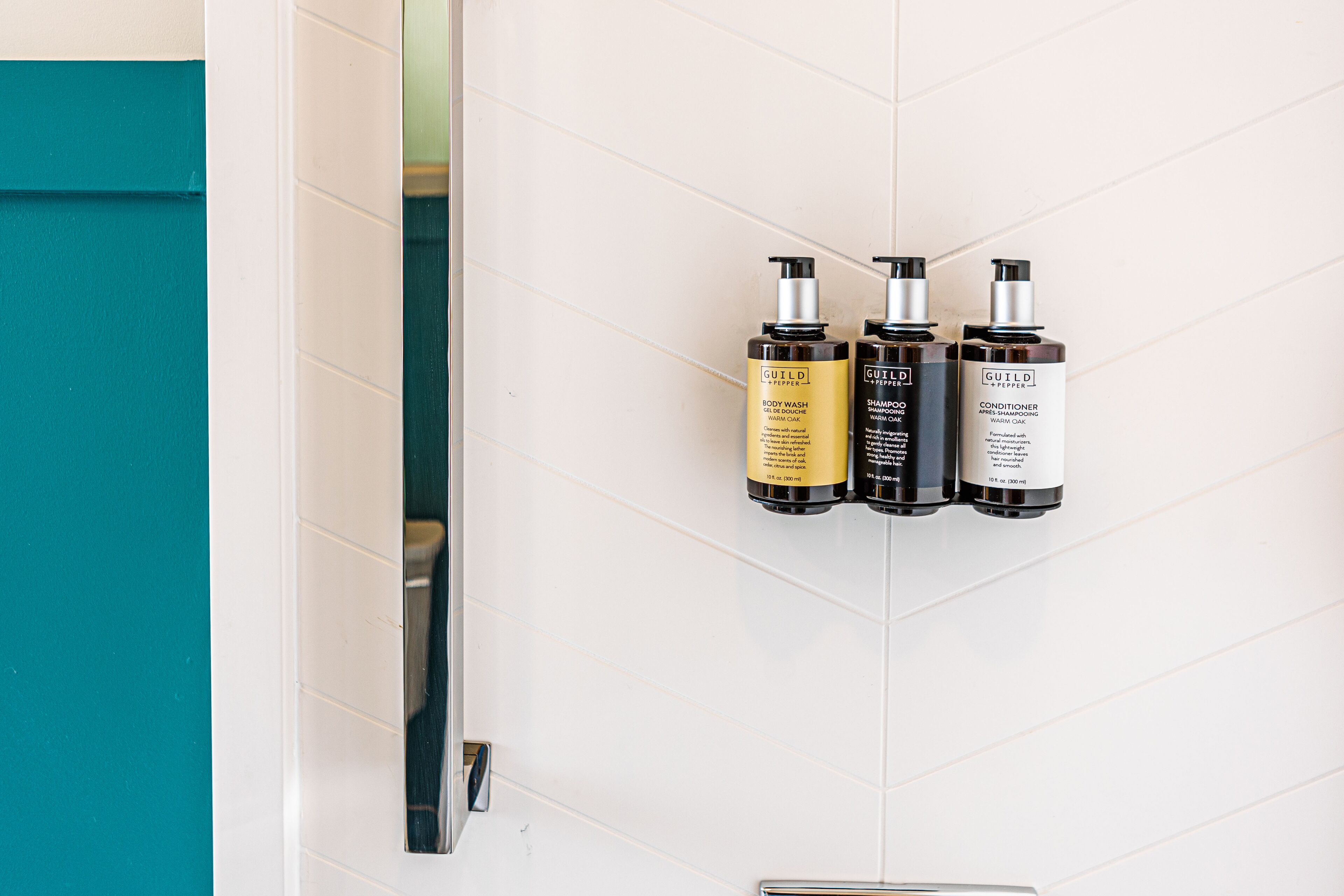 Three bottles of Guild L+Pepper body wash, shampoo, and conditioner on a white tiled wall.