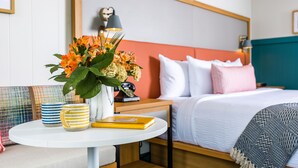 A neatly arranged hotel room with a bed, bedside table, and a small round table with a vase of flowers, a book, and striped mugs. - Freebird Motor Lodge by Reverie Boutique Collection (West Yarmouth)