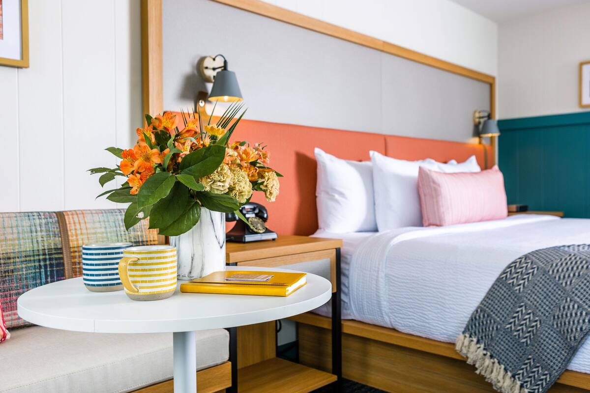 Quarto de hotel bem arrumado, com cama, mesa de cabeceira e uma pequena mesa redonda com um vaso de flores, um livro e canecas listradas.