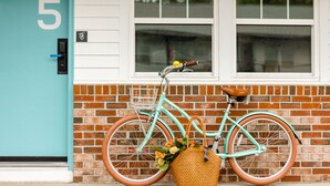 A teal bicycle with a basket and a wicker bag leaning against a brick wall with a white door marked with the number 5. - Freebird Motor Lodge by Reverie Boutique Collection (West Yarmouth)