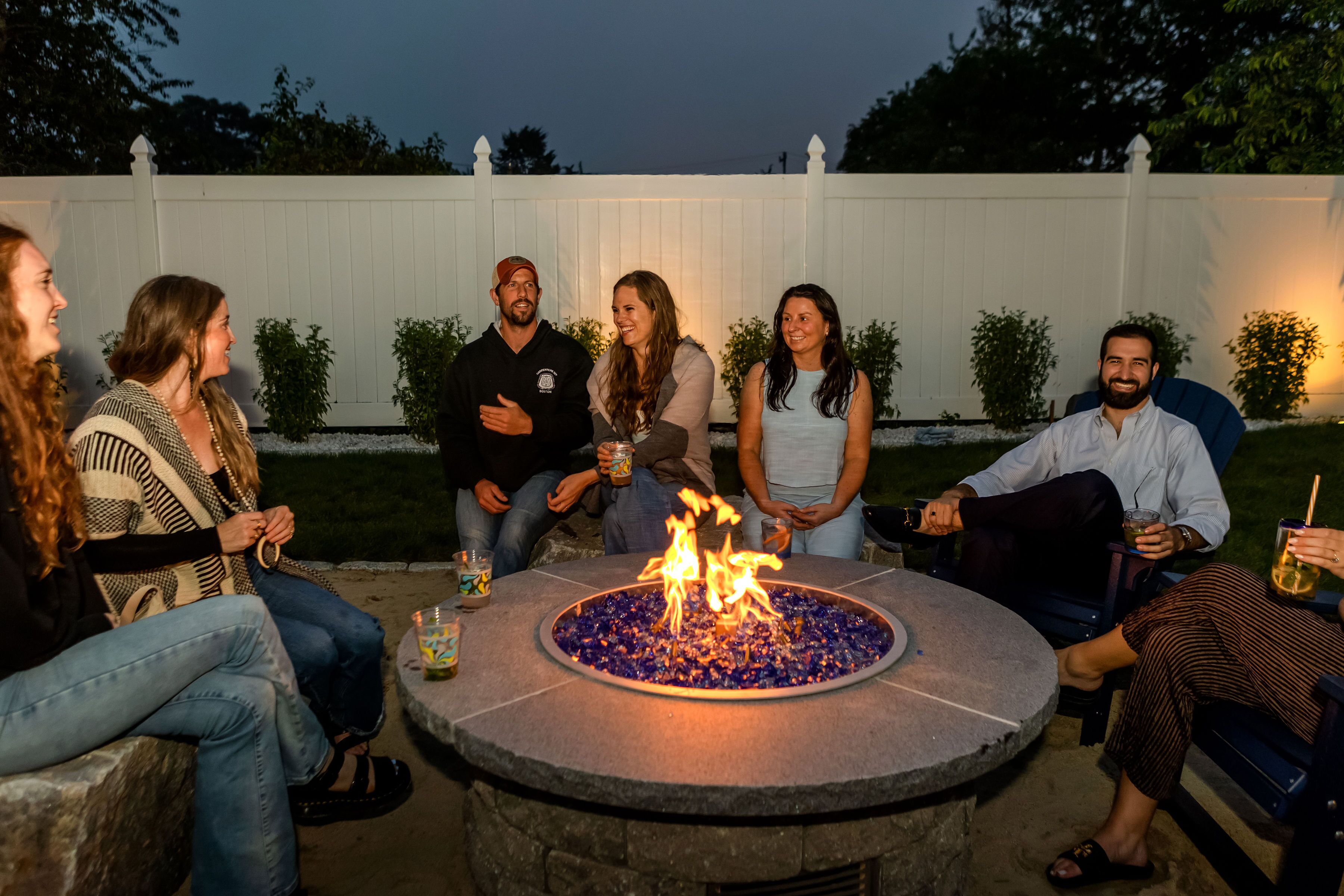 A group of people seated around a stone fire pit with a white fence in the background.