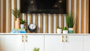 A modern living room with a mounted TV, wooden panel wall, books, a clock, and decorative plants. - Freebird Motor Lodge by Reverie Boutique Collection (West Yarmouth)