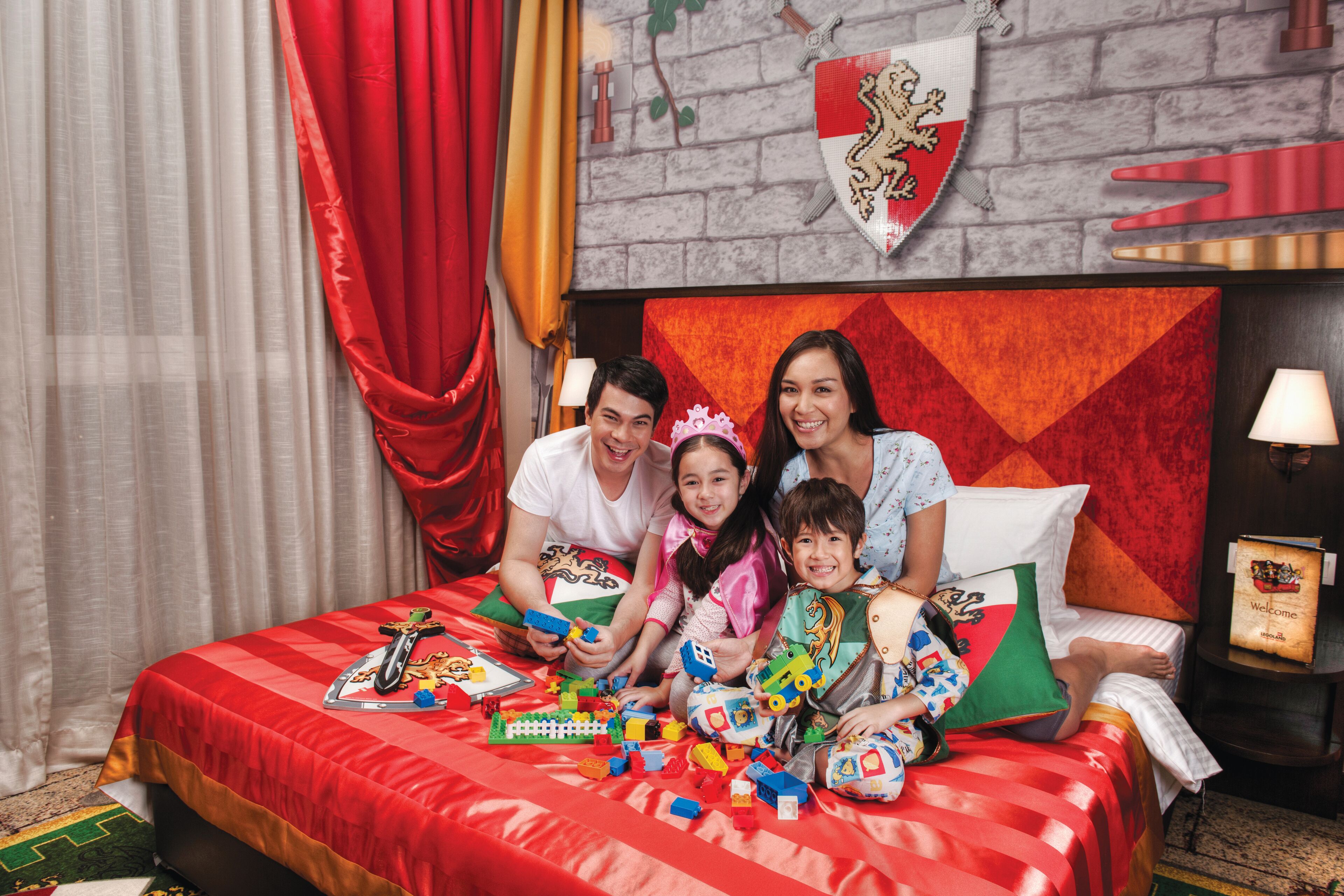 A family on a bed with a red bedspread, toys, and a shield with a lion emblem on the wall.