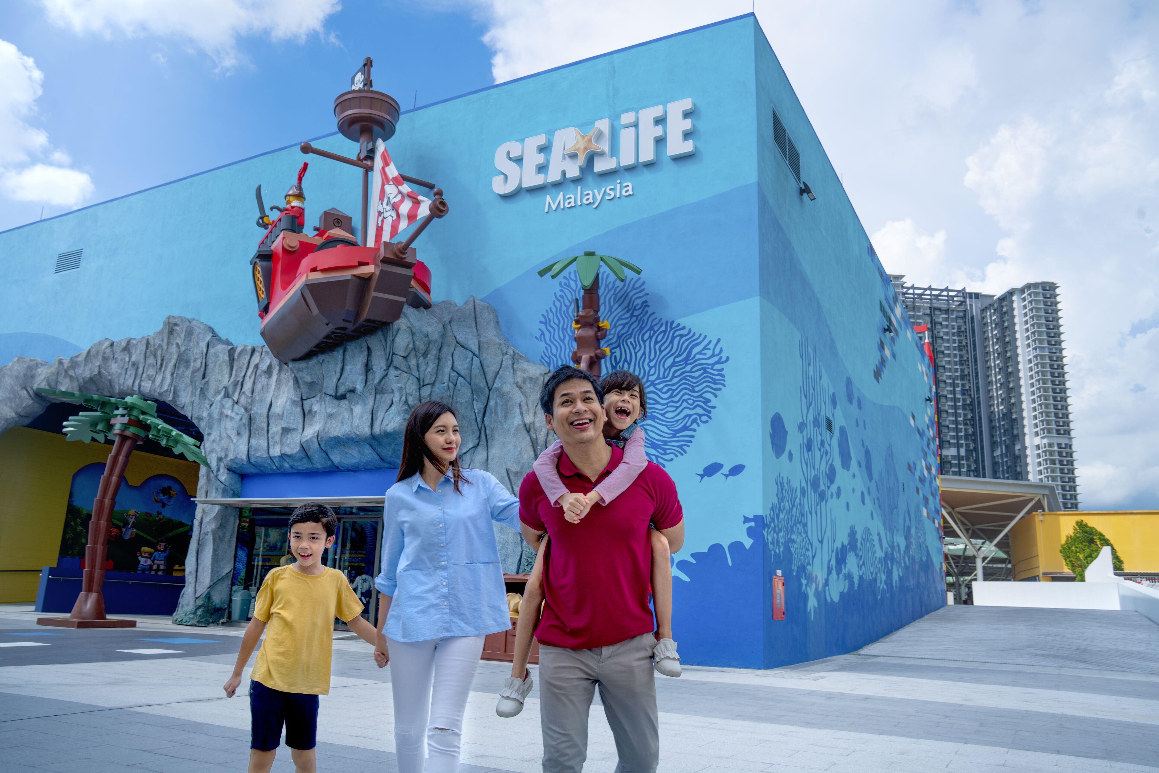 Eine Familie geht vor einem Gebäude entlang, das eine große Wandmalerei eines Schiffes zeigt. Auf der Wandmalerei steht außerdem 'SEA LIFE Malaysia'.