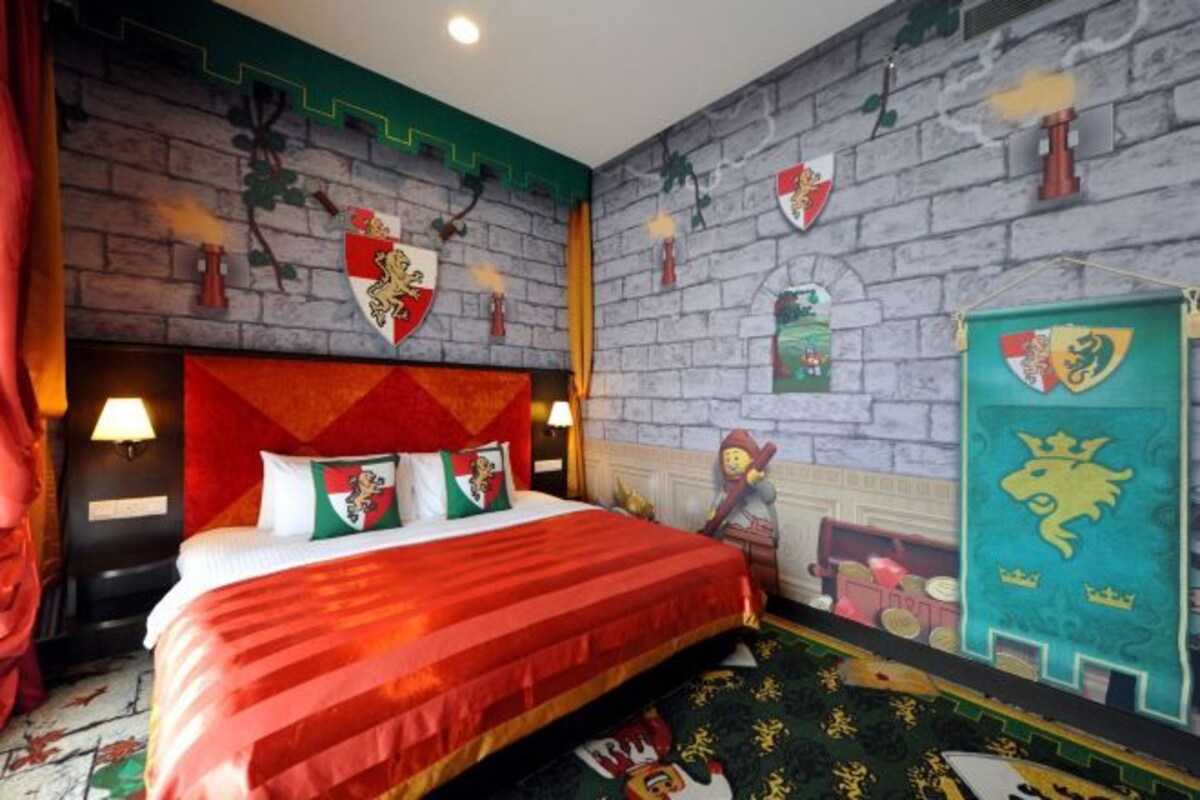 Una habitación de hotel con una cama grande, un cabecero con diseño rojo y blanco, y una pared decorada con varios escudos heráldicos.