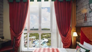 A hotel room with a large window, red curtains, a bed with striped bedding, a pillow with a coat of arms, and a view of a water park.