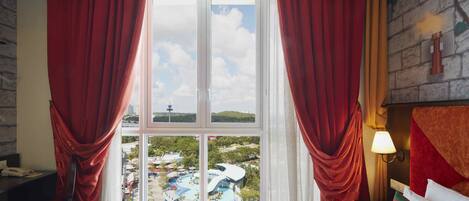 A hotel room with a large window, red curtains, a bed with striped bedding, a pillow with a coat of arms, and a view of a water park.