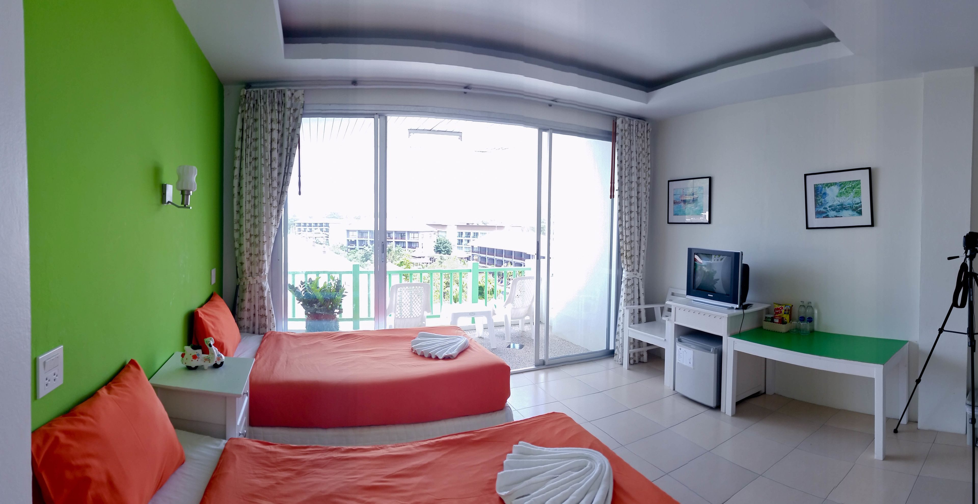 A hotel room with two beds, a TV, a desk with a computer, and a balcony with a view of buildings.