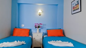 A room with two beds, each with an orange pillow and a blue bedspread. There is a nightstand with a vase of flowers and a wall-mounted light.