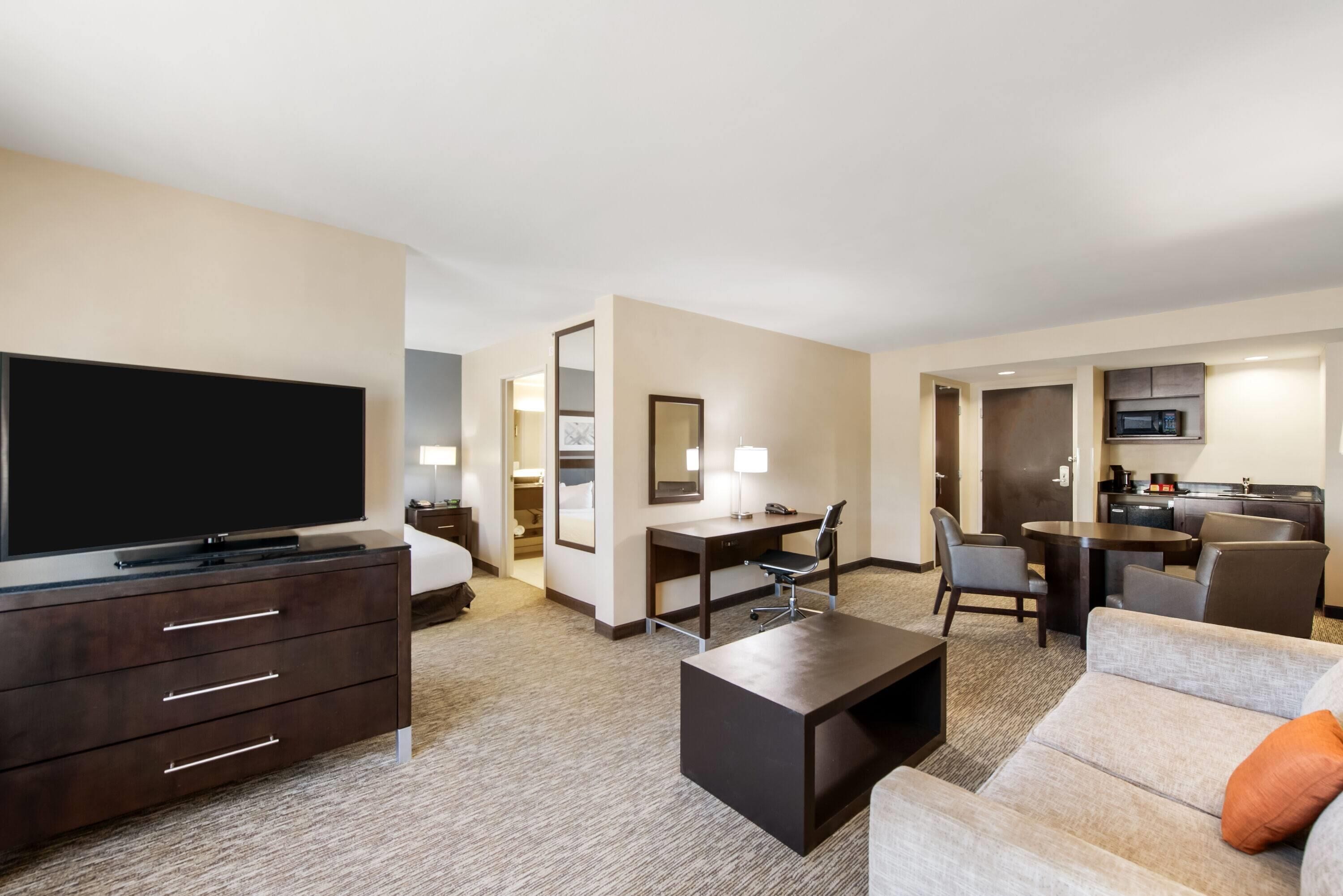 A hotel room with a TV, a desk, a chair, a sofa, and a kitchen area.