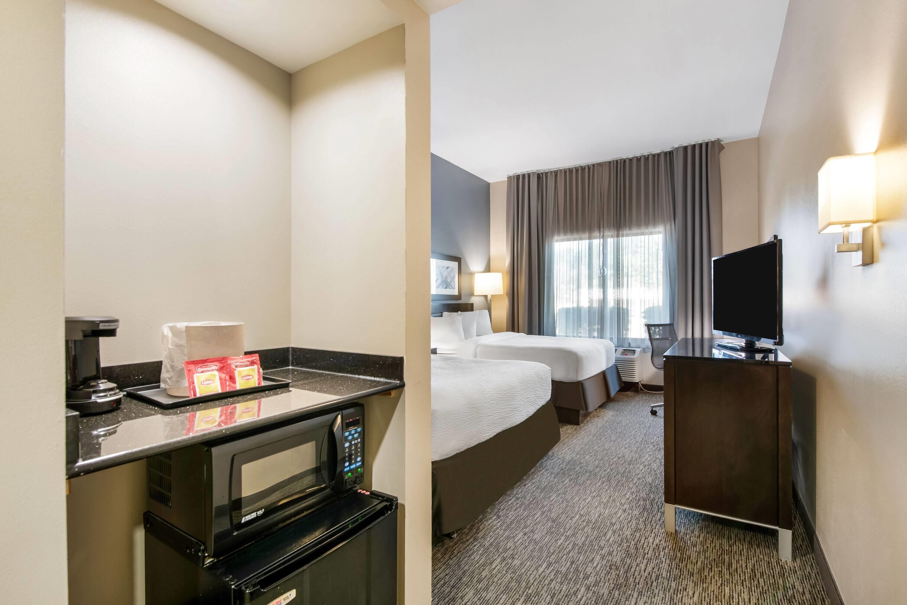 Quarto de hotel com cozinha compacta, duas camas, televisão e uma janela grande com cortinas.