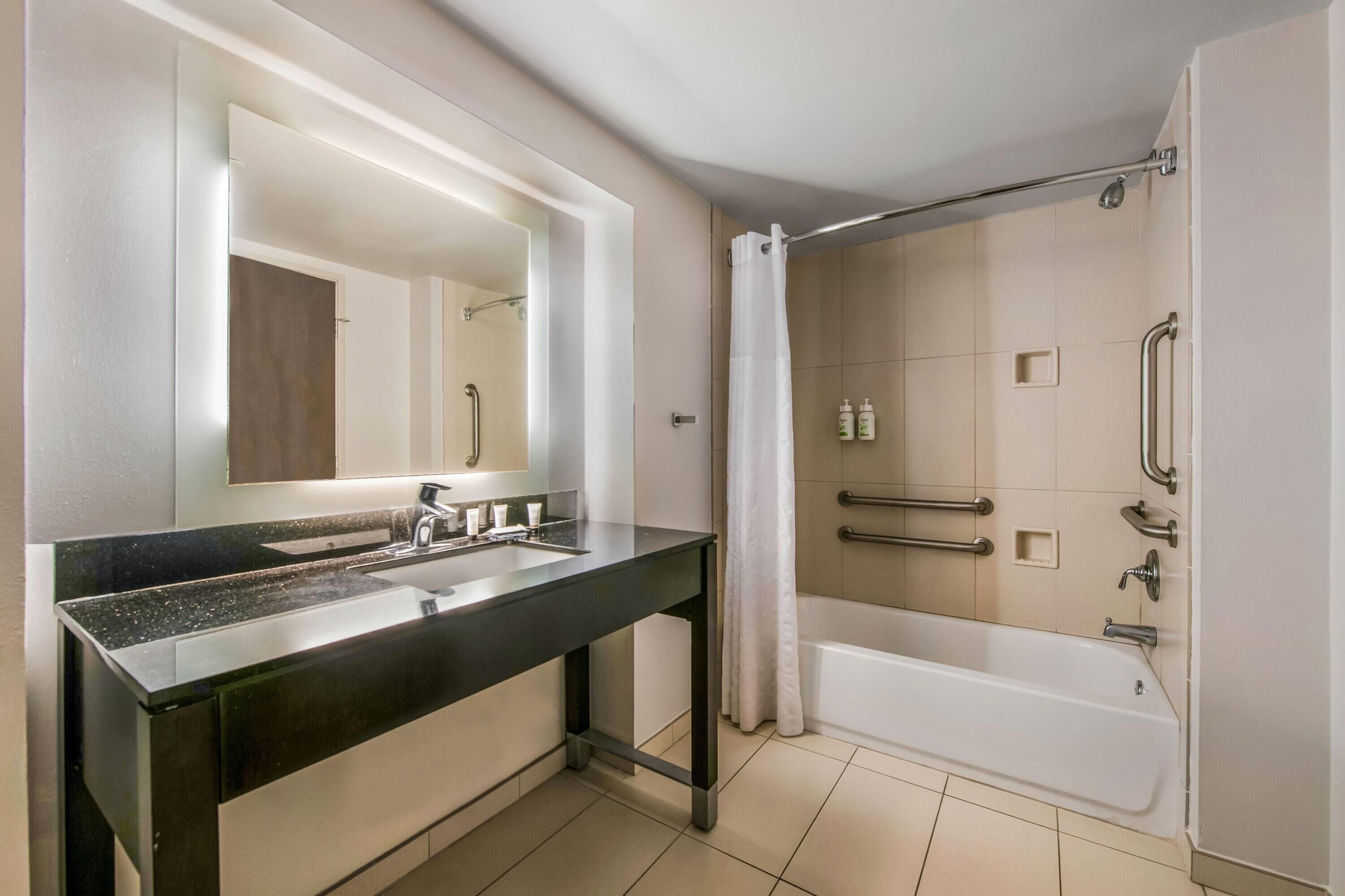 A modern bathroom with a double sink vanity, a large mirror, a bathtub with a shower curtain, and handrails.
