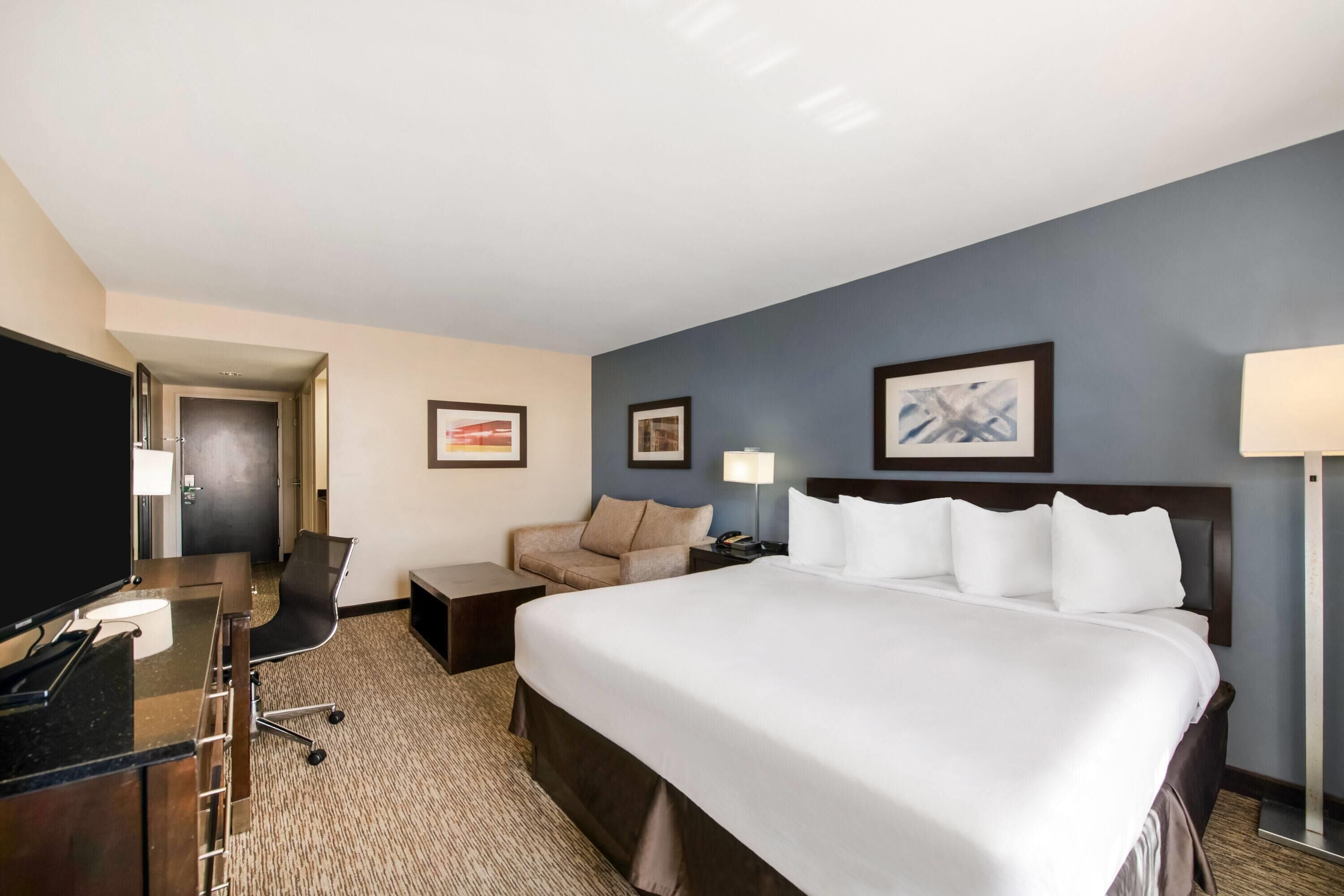 Ein Hotelzimmer mit einem großen Bett, einem Schreibtisch mit Computer, einem Sessel, einem Fernseher und gerahmten Bildern an der Wand.