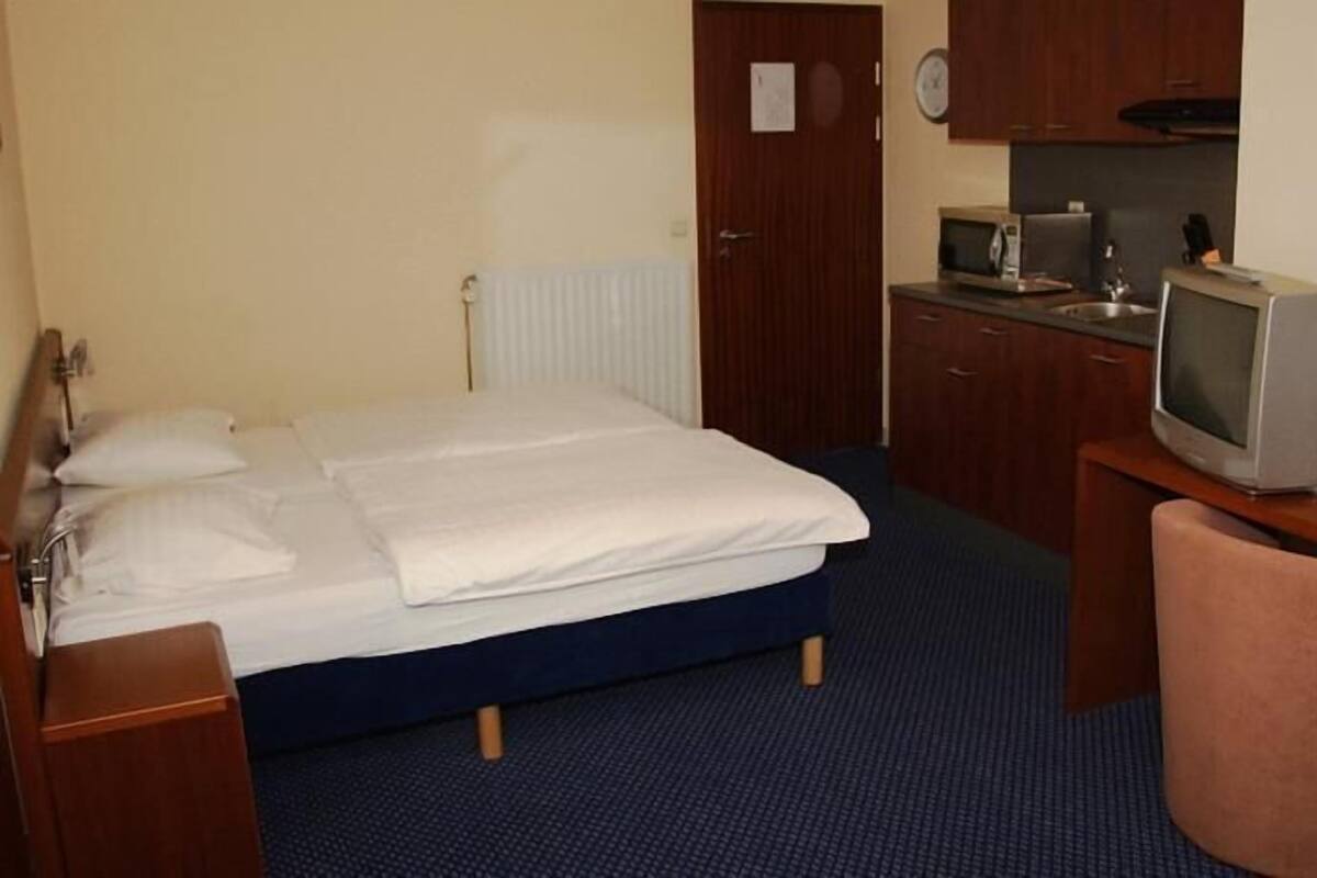 A hotel room with a bed, a desk with a television, a chair, and a kitchenette area.