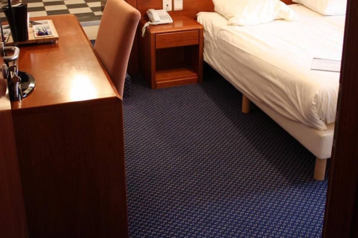 A hotel room with a bed, a desk, a chair, a nightstand, a lamp, and a painting on the wall.