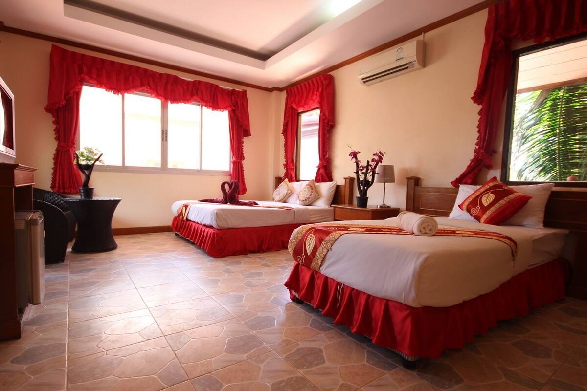 A hotel room with two beds, red curtains, a potted plant, a desk, and a window with a view of greenery.