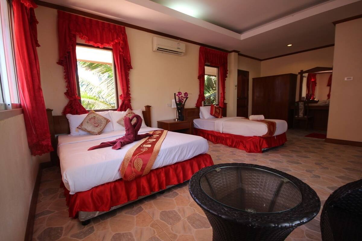A hotel room with two beds, red curtains, a stone-tiled floor, a glass table, and a trash bin.