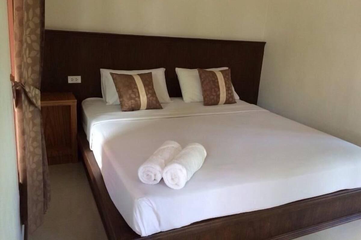 A neatly made bed with white linens and two rolled towels, a wooden headboard, and a nightstand with a lamp.