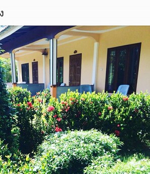 A covered outdoor area with a yellow building, a blue chair, and a garden with red flowers. - Phu Lanta Life (Ko Lanta)