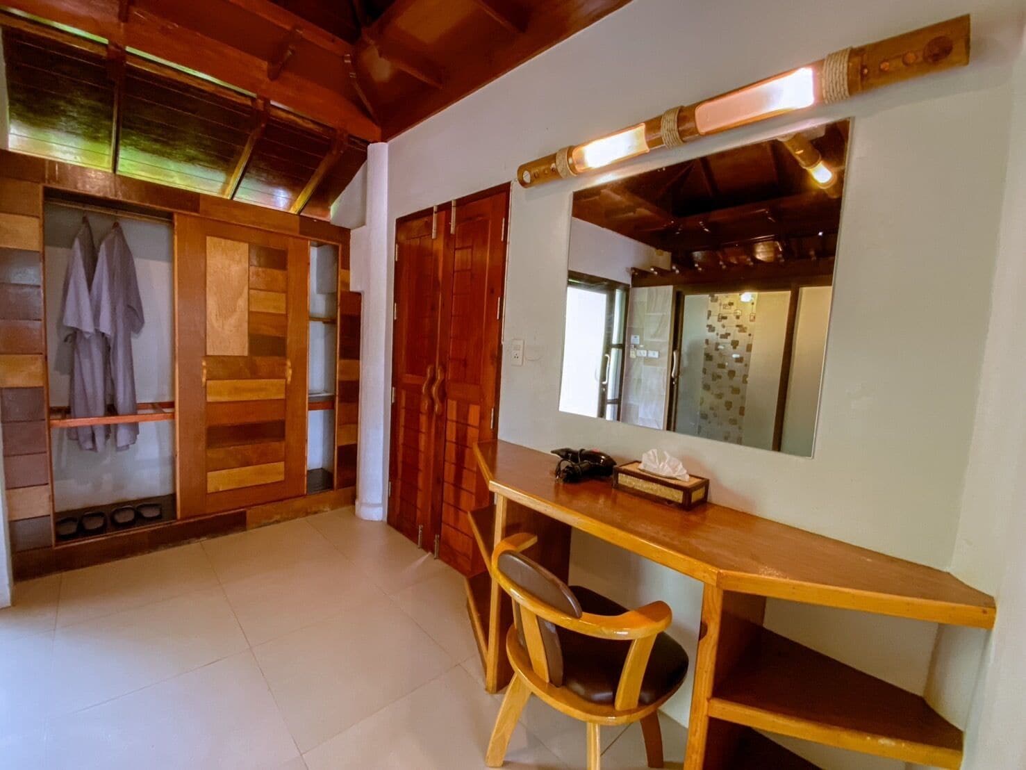 Una habitación con un escritorio de madera, una silla y un espejo encima. Hay un armario con puertas correderas y un estante con zapatos.