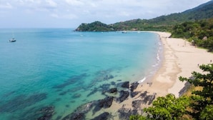 Une plage côtière aux eaux turquoise et limpides, bordée d’une côte rocheuse, d’où l’on aperçoit une petite île au loin.