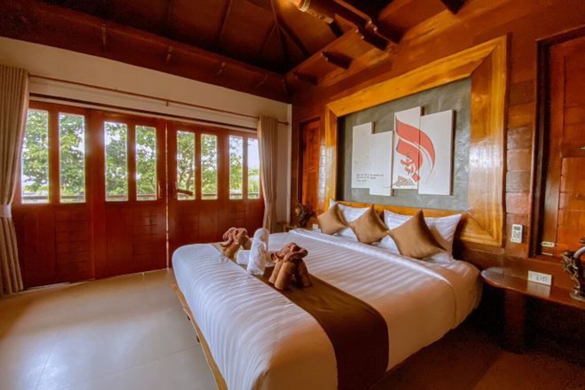 Ruime, smaakvolle slaapkamer met houten plafond, een groot bed met veel kussens en uitzicht op groen door de ramen.