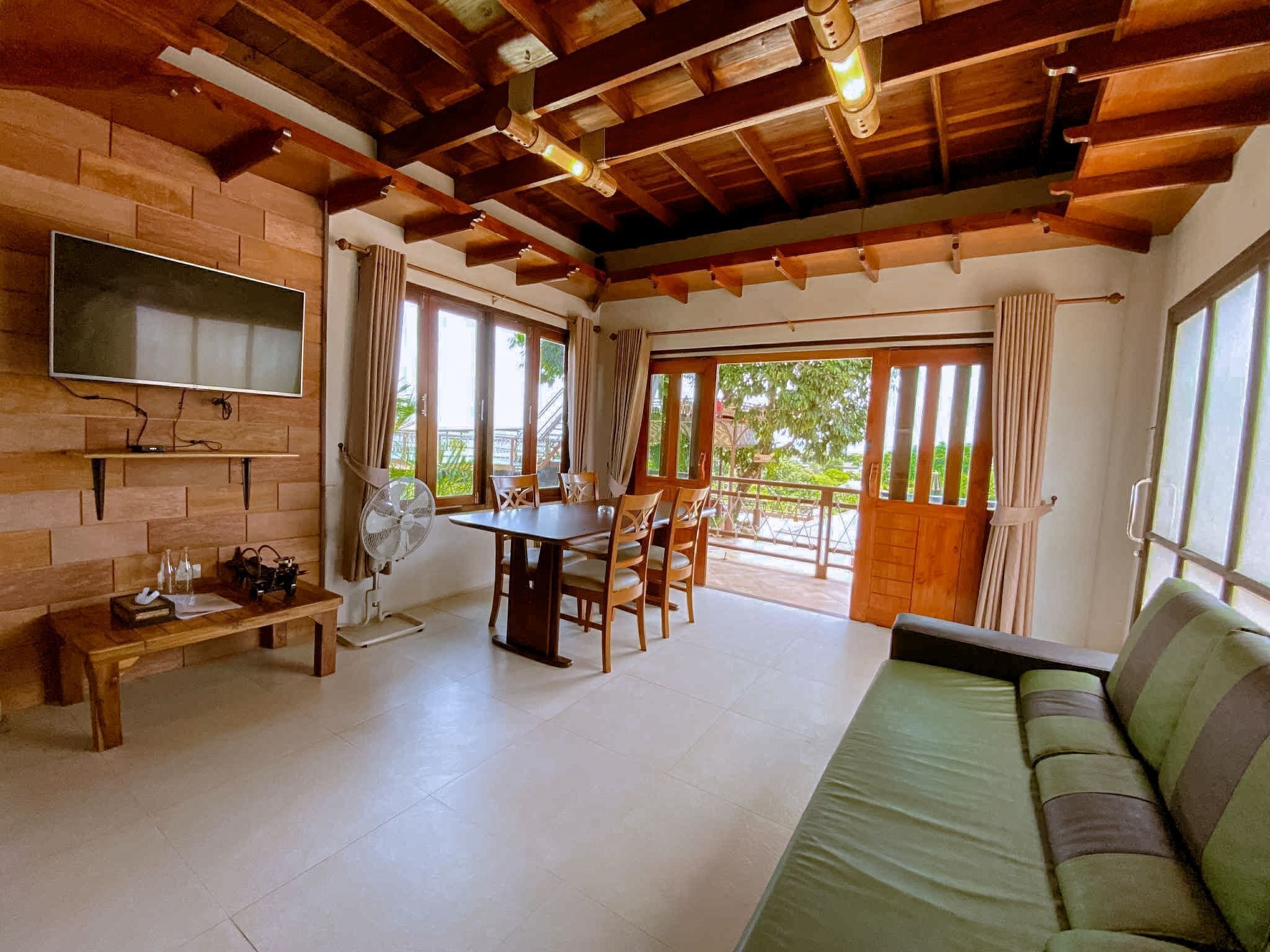 Una habitación acogedora con techo de madera, un televisor montado en la pared, una mesa de comedor con sillas, un sofá y ventanas amplias con cortinas.