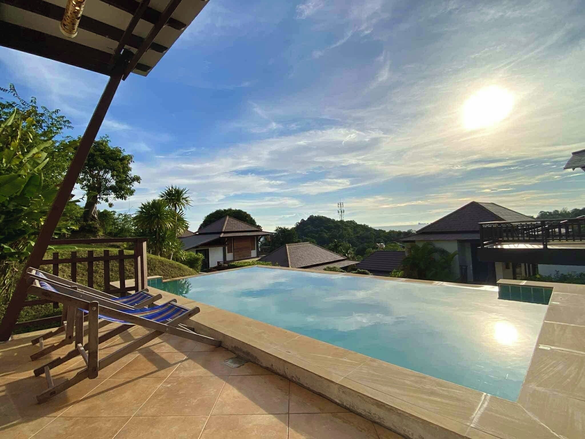 A swimming pool with a wooden deck and lounge chairs, surrounded by houses and greenery.