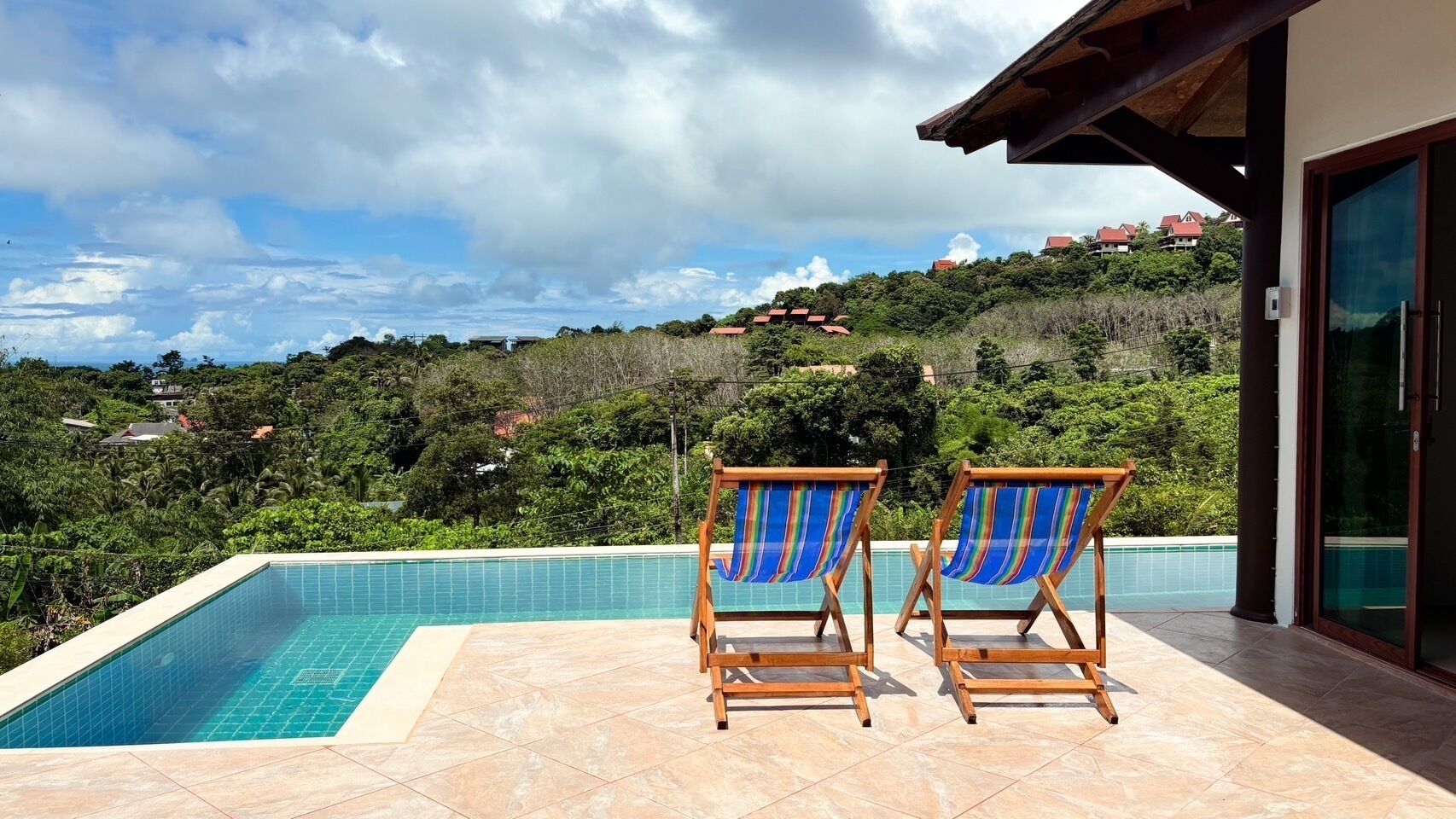 Una piscina con dos sillones de madera y toallas azules y blancas a rayas, rodeada por un suelo de baldosas.