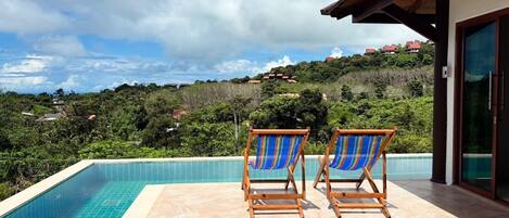 Una piscina con dos sillones de madera y toallas azules y blancas a rayas, rodeada por un suelo de baldosas.