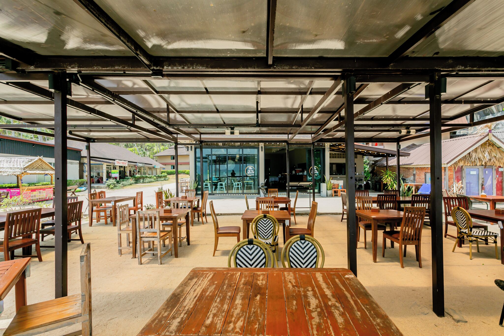 An outdoor cafe with wooden tables and chairs, a covered seating area, and a view of the street and buildings in the background.