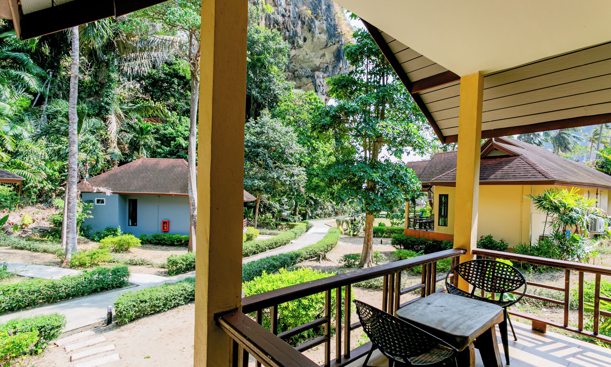 A covered outdoor area with a table and chairs, surrounded by lush greenery and a pathway leading to buildings.