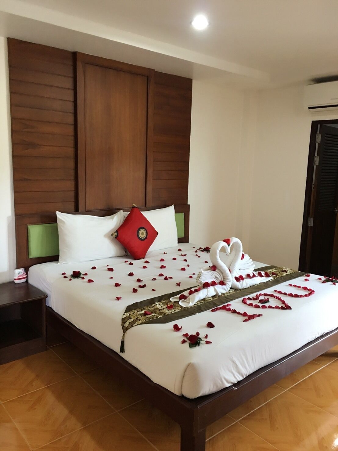 A neatly made bed with a swan-shaped towel arrangement and rose petals, a red pillow, and a green headboard.