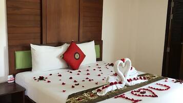 A neatly made bed with a swan-shaped towel arrangement and rose petals, a red pillow, and a green headboard.