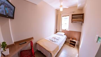 A hotel room with a bed, a desk, a chair, a TV, a ceiling fan, a window with curtains, and a small shelf.